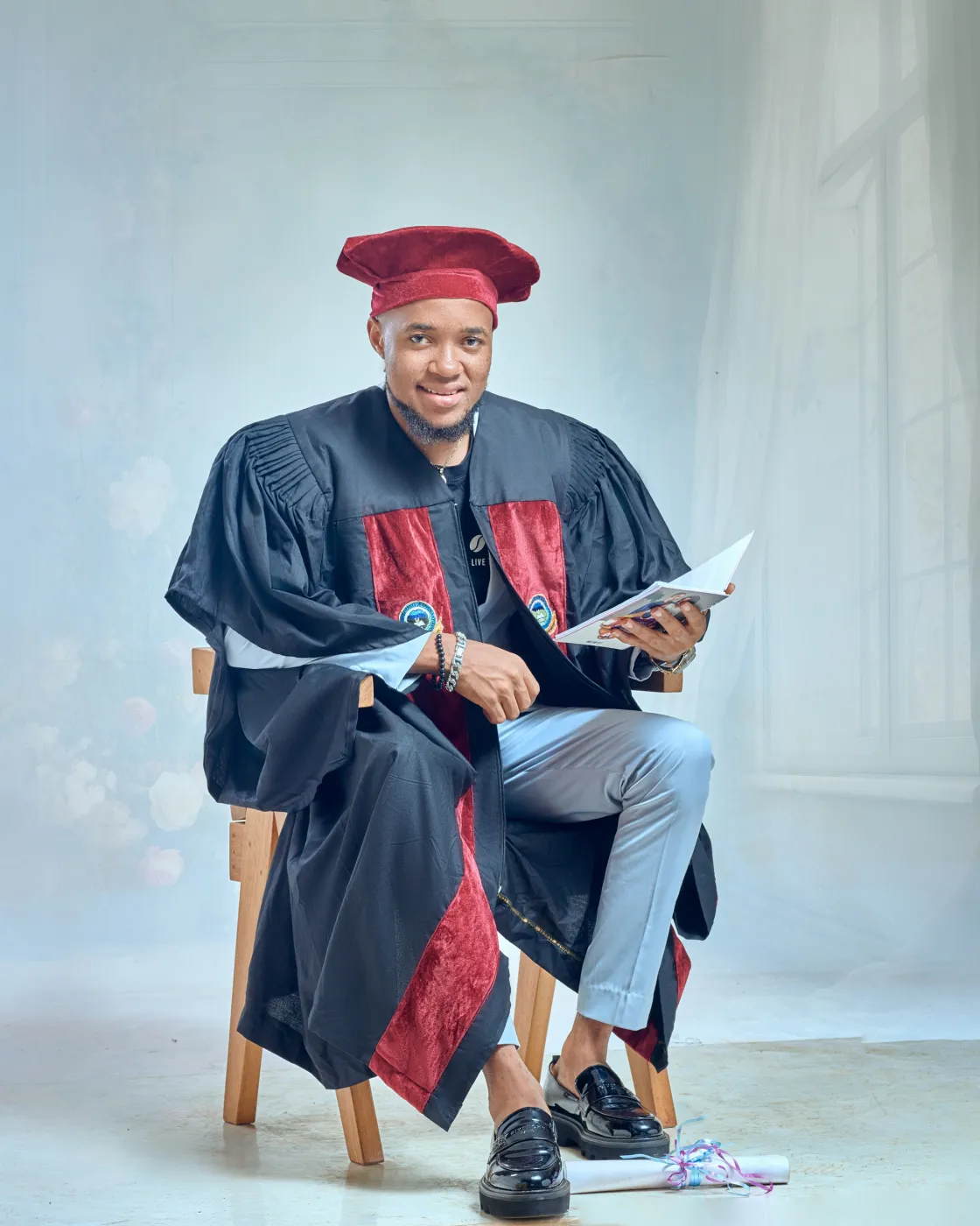 Nsom Emmanuel seated in a black and red graduation gown and cap, holding a book